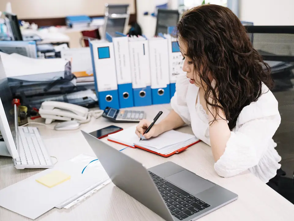 Office worker taking notes at a desk with a laptop, a desk phone and binders